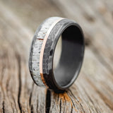 Shown here is "Tanner", a custom, handcrafted hammered men's wedding ring featuring antler and 14K rose gold inlay, shown here on a fire-treated black zirconium band, upright facing left. Additional inlay options are available upon request.