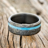 Shown here is "Vertigo", a custom, handcrafted men's wedding ring featuring an offset turquoise inlay, shown here on a fire-treated black zirconium band with a floral pattern engraving, laying flat. Additional inlay options are available upon request.