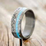 Shown here is "Vertigo", a custom, handcrafted men's wedding ring featuring an offset turquoise inlay, shown here on a fire-treated black zirconium band with a floral pattern engraving, upright facing left. Additional inlay options are available upon request.