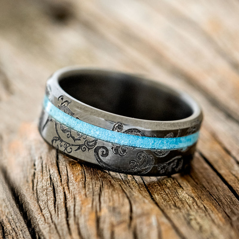 Shown here is "Vertigo", a custom, handcrafted men's wedding ring featuring an offset turquoise inlay, shown here on a fire-treated black zirconium band with a floral pattern engraving, tilted left. Additional inlay options are available upon request.