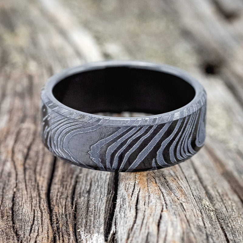 Shown here is a handcrafted men's wedding ring shown featuring a solid band with a woodgrain pattern finish, laying flat.