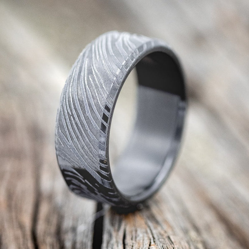 Shown here is a handcrafted men's wedding ring featuring a solid black zirconium wedding band with a unique woodgrain pattern etched into the metal, upright facing left. Additional inlay options are available upon request.