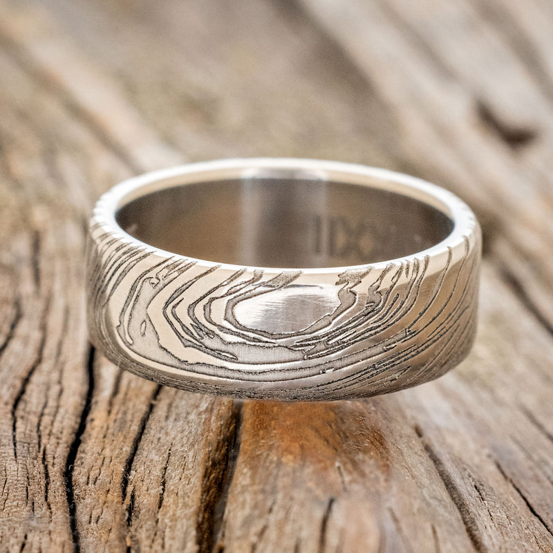 Shown here is a handcrafted men's wedding ring shown featuring a solid band with a woodgrain pattern finish, laying flat.