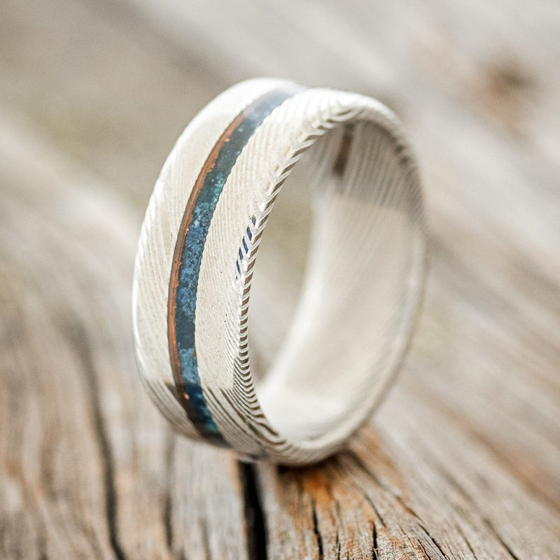 Shown here is "Nirvana", a custom, handcrafted men's wedding ring featuring a centered patina copper inlay on a Damascus steel band, upright facing left. Additional inlay options are available upon request.