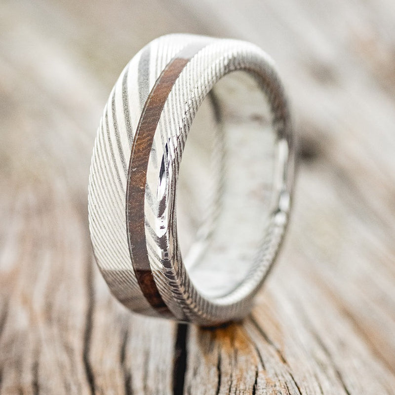 Shown here is "Vertigo", a custom, handcrafted antler-lined men's wedding ring featuring an ironwood inlay on a Damascus steel band, upright facing left. Additional inlay options are available upon request.