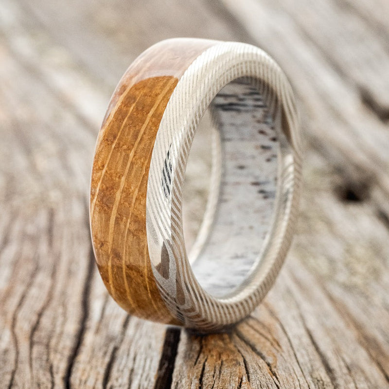 Shown here is "Ezra", a custom, handcrafted men's wedding ring featuring a whiskey barrel wood overlay and an antler lined titanium band, upright facing left. Additional inlay and lining options are available upon request.