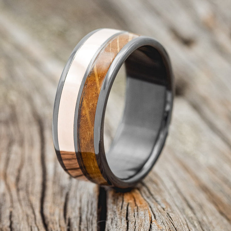 Shown here is "Dyad", a custom, handcrafted men's wedding ring featuring 2 channels with 14K rose gold and whiskey barrel oak inlays, shown here set on a fire-treated black zirconium band, upright facing left. Additional inlay options are available upon request.