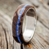 Shown here is "Remmy", a custom, handcrafted men's wedding ring featuring an ironwood overlay and an offset lapis lazuli inlay, shown here on a titanium band, upright facing left. Additional inlay options are available upon request.