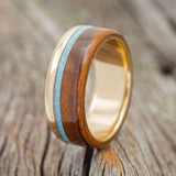 Shown here is "Ezra", a custom, handcrafted wedding band featuring a 14K yellow gold band with turquoise and ironwood, upright facing left. Additional inlay options are available upon request.