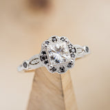Shown here is "Eileen", a vintage-style moissanite women's engagement ring with black diamond accents, on stand front facing. Many other center stone options are available upon request.