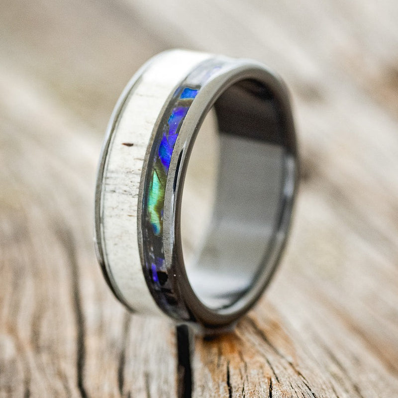 Shown here is "Raptor", a custom, handcrafted men's wedding ring featuring a Paua shell & antler inlay on a fire-treated black zirconium band, upright facing left. Additional inlay options are available upon request.