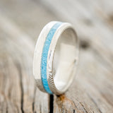 Shown here is "Vertigo", a custom, handcrafted narrow men's wedding ring featuring a turquoise inlay on a Damascus steel band, upright facing left. Additional inlay options are available upon request.