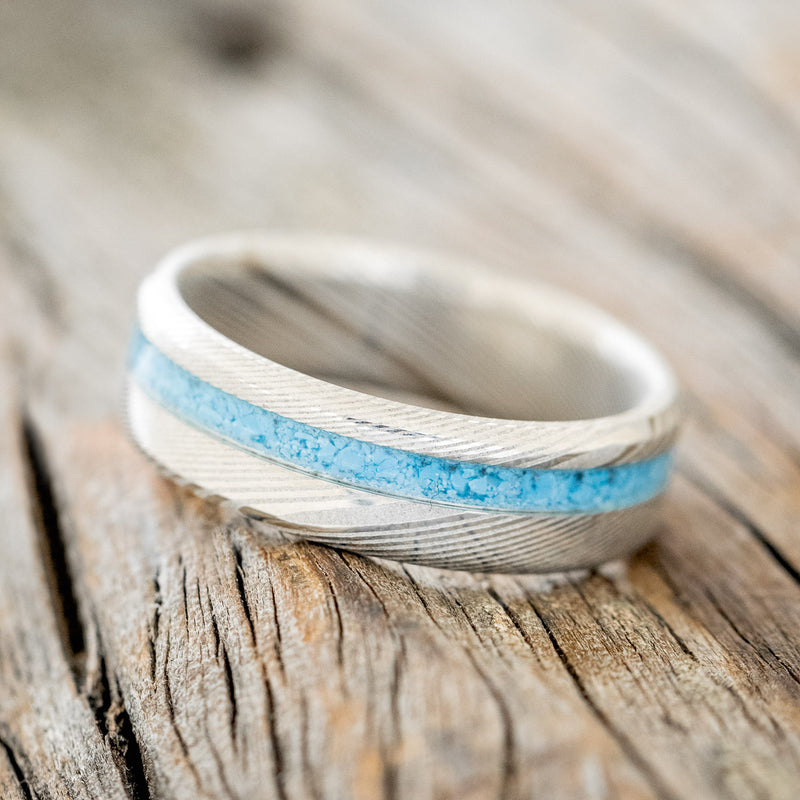 Shown here is "Vertigo", a custom, handcrafted narrow men's wedding ring featuring a turquoise inlay on a Damascus steel band, tilted left. Additional inlay options are available upon request.