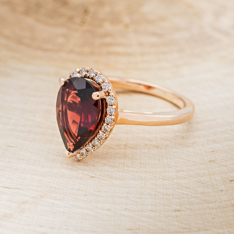 Shown here is a pear-shaped garnet women's engagement ring with a diamond halo, facing left. Many other center stone options are available upon request.