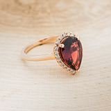 Shown here is a pear-shaped garnet women's engagement ring with a diamond halo, facing right. Many other center stone options are available upon request.