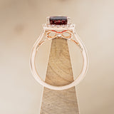 Shown here is a pear-shaped garnet women's engagement ring with a diamond halo, side view on stand. Many other center stone options are available upon request.