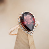 Shown here is a pear-shaped garnet women's engagement ring with a diamond halo, on stand facing slightly right. Many other center stone options are available upon request. 
