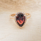 Shown here is a pear-shaped garnet women's engagement ring with a diamond halo, front facing. Many other center stone options are available upon request.