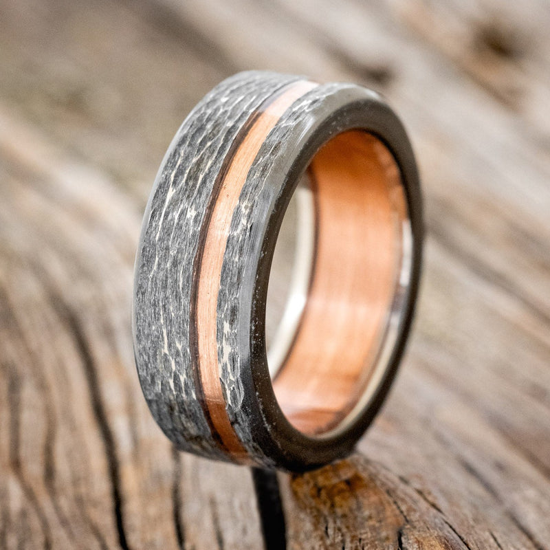 Shown here is "Vertigo", a custom, handcrafted hammered men's wedding ring featuring a rustic copper lining and inlay on a fire-treated black zirconium band, upright facing left. Additional inlay options are available upon request.