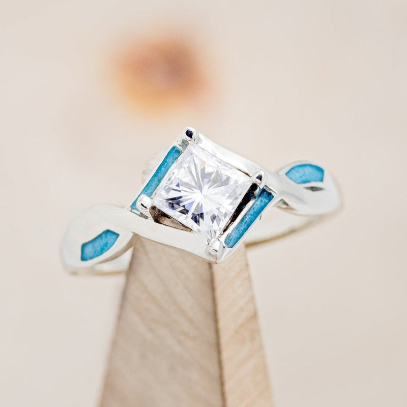 Shown here is "Adamas", a prong-style moissanite women's engagement ring with turquoise inlays, on stand front facing. Many other center stone options are available upon request. 