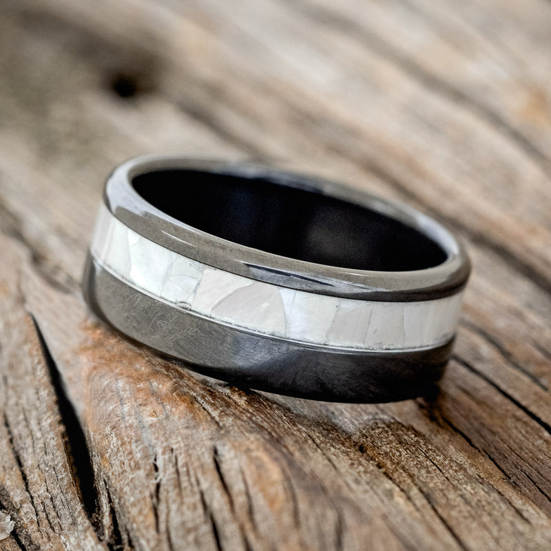 Shown here is "Castor", a custom, handcrafted men's wedding ring featuring a mother of pearl inlay on a fire-treated black zirconium band, tilted left. Additional inlay options are available upon request.