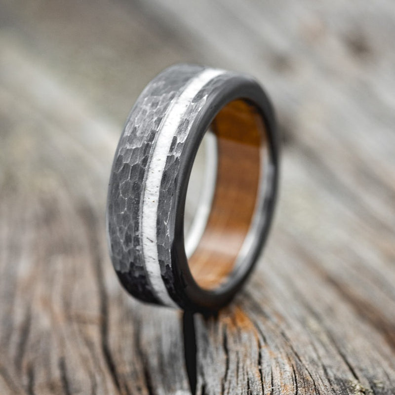 Shown here is "Vertigo", a handcrafted men's wedding ring shown with a fire-treated black zirconium hammered wedding band with whiskey barrel lining and elk antler inlay, upright facing left. Additional inlay options are available upon request.