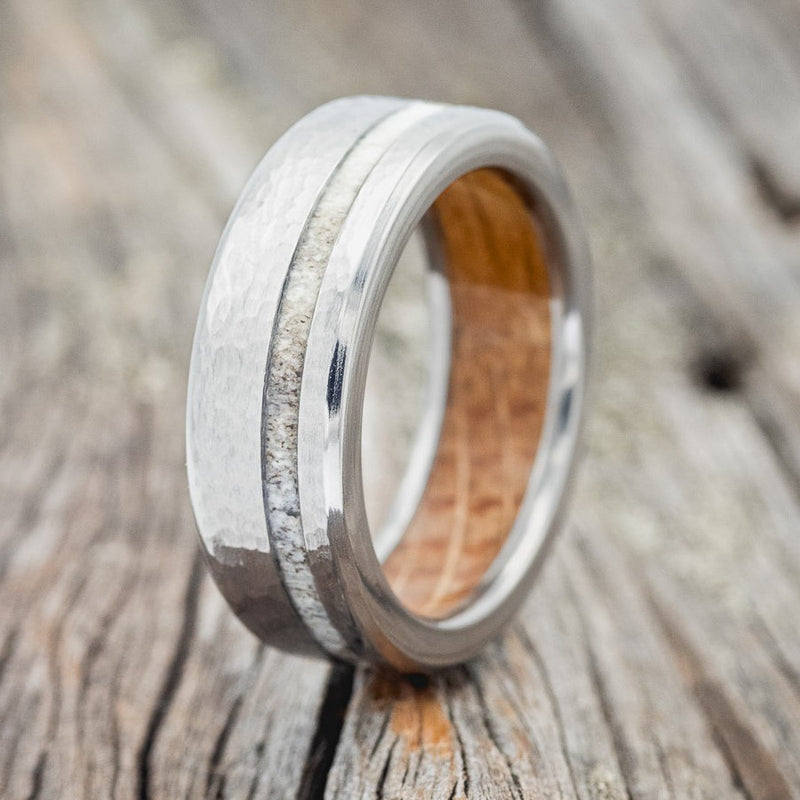 Shown here is "Vertigo", a handcrafted men's wedding ring featuring a whiskey barrel lining, an antler inlay, and a hammered finish, upright facing left. Additional inlay options are available upon request.