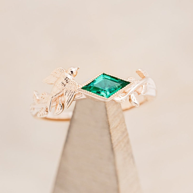 Shown here is "Sparrow", a lab-created emerald women's engagement ring with a sparrow embellishment, on stand front facing. Many other center stone options are available upon request.  