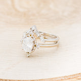 Shown here is "Treva", an oval moissanite women's engagement ring set with diamond accents and a "Melody" tracer, facing left. Many other center stone options are available upon request.