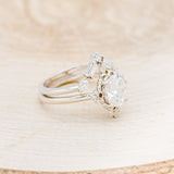 Shown here is "Treva", an oval moissanite women's engagement ring set with diamond accents and a "Melody" tracer, facing right. Many other center stone options are available upon request.