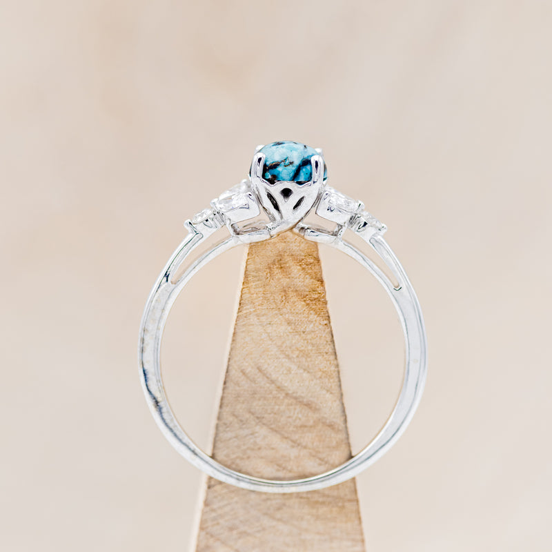 Shown here is "Blossom", a turquoise women's engagement ring with leaf-shaped diamond accents, side view on stand. Many other center stone options are available upon request.