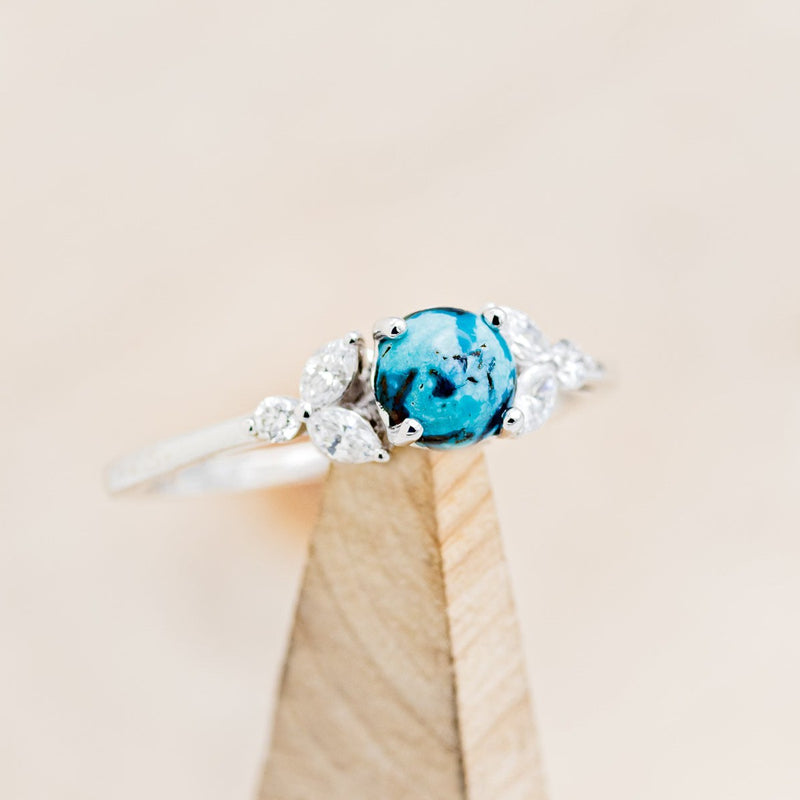 Shown here is "Blossom", a turquoise women's engagement ring with leaf-shaped diamond accents, on stand facing slightly right. Many other center stone options are available upon request. 