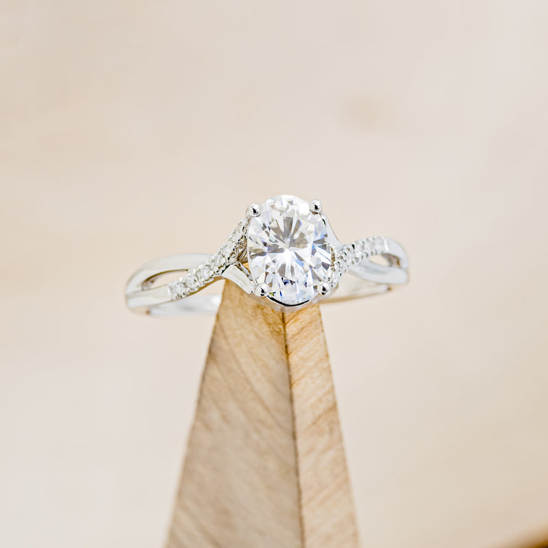 Shown her is "Roslyn", an oval moissanite women's engagement ring with diamond accents, on stand front facing. Many other center stone options are available upon request.