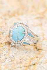 Shown here is  The "KB", a halo-style oval turquoise women's engagement ring with delicate and ornate details and is available with many center stone options. shown here with two diamond and turquoise inlays tracers.