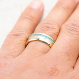 Shown here, Vertigo, a custom, handcrafted men's wedding ring featuring an offset turquoise inlay, shown here on a yellow gold band, on hand. Additional inlay options are available upon request.