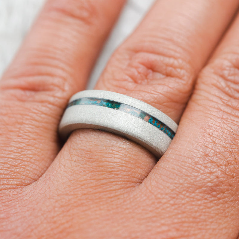 Shown here, Vertigo, a custom, handcrafted men's wedding ring featuring a sandblasted titanium base, with an offset copper patina inlay and a turquoise lining, on hand. Additional inlay options are available upon request.